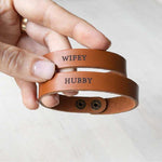 two leather matching couples bracelets in tan held by a female Caucasian hand. the matching bracelets are engraved with hubby and wifey for a husband and wife third anniversary gift.